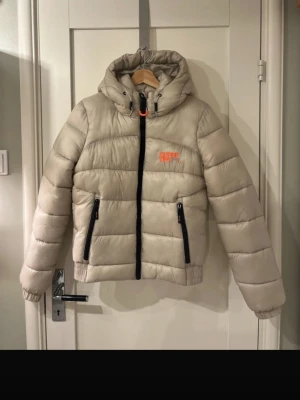 Beige pufferjacka från Superdry - Beige pufferjacka från Superdry med svart dragkedja och detaljer. Jackan har huva med snörning, två stora fickor med dragkedja och orange Superdry-logga på sopbröstet. Perfekt för kalla dagar och riktigt snygg!