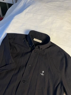 Svart kortärmad skjorta The Blue Uniform - Svart skjorta från The Blue Uniform i storlek S. Skjortan har korta ärmar, klassisk krage med knappar och en vit broderad ankare på bröstet. Tillverkad i mjuk bomull och har en clean, stilren look.
