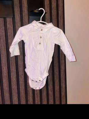 Vit bodyskjorta från Newbie storlek 68 - Klassisk vit bodyskjorta från Newbie med lång ärm, krage och knappar framtill. Tillverkad i mjuk bomull och har praktiska tryckknappar i grenen. Perfekt för en clean och stilren look till de minsta. Storlek 68