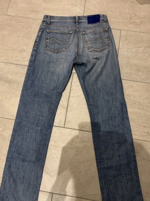 Blå jeans från Jacob Cohen, storlek 32 - Säljer ett par klassiska blå jeans från Jacob Cohen i storlek 32. Jeansen har raka ben, fem fickor och snygga kontrastsömmar. Märkespatch i blå sammet baktill och slitning på vänster lår för en cool look. Perfekta till vardagsoutfiten.