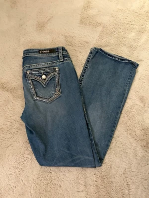 Blå Vigoss jeans med bootcut - Säljer ett par blå jeans från Vigoss i modellen Slim Boot och Heritage Fit. Jeansen har snygga kontrastsömmar, dekorativa fickor med blingdetaljer och klassisk femficksdesign. Perfekt passform med bootcut och låg midja. Materialet är slitstarkt jeans. Liten  fläck som inte verkar gå bort men absolut inget som syns. 