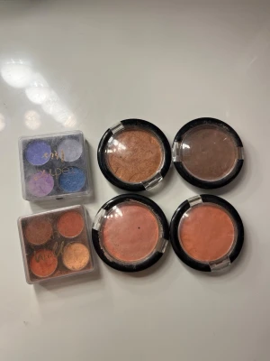 Mixade rouge och ögonskuggspaletter - Säljer fyra runda rouge i persika, rosa, brun och koppar samt två fyrfärgade ögonskuggspaletter. Ena paletten har lila, blå och silver toner, den andra har varma nyanser som orange, koppar och guld. Alla produkter i praktiska plastförpackningar. De är använda ett par gånger.