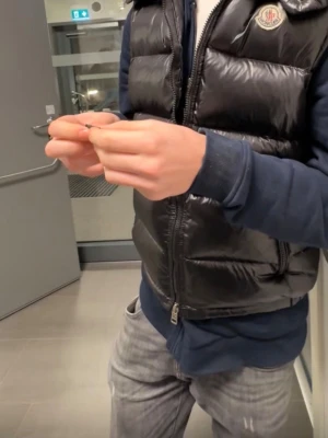 Svart dunväst från Moncler - Säljer en svart, glansig dunväst från Moncler med klassisk logga på bröstet. Västen har dragkedja framtill, två fickor med dragkedja och en hög krage. Perfekt för lager-på-lager och riktigt snygg till streetwear-stil.
