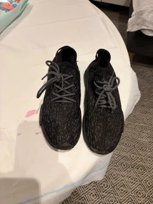 Svarta Adidas NMD sneakers - Säljer ett par svarta Adidas NMD sneakers med stickad ovandel och coola grå snören. Skorna har diskret mönster i svart och grått, samt en röd detalj på hälen. Sulan är i svart och ger grym komfort till vardags. Perfekta för dig som gillar streetwear och snygg design.