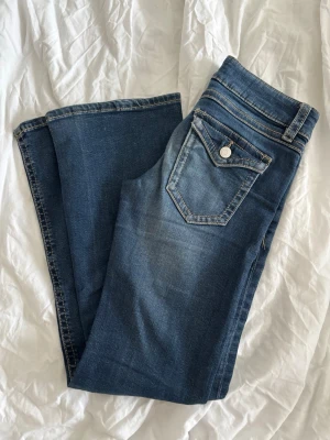 Blå bootcut jeans med fickdetaljer - ‼️PRIS KAN DISKUTERAS‼️STORLEK 158! Säljer ett par klassiska blå jeans från Ginatricot med bootcut passform eftersom de är för små för mig. Jeansen har snygga kontrastsömmar, fem fickor och dekorativa knappar på bakfickorna. Materialet är mjukt denim i bomull med lite stretch. 