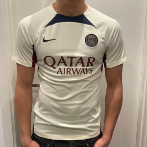 PSG matchtröja Nike vit/röd - Säljer en Paris Saint-Germain matchtröja från Nike i crémevitt med blå/vinröda detaljer och logga. Tröjan har röda inslag vid axlarna. Materialet är syntet och perfekt för fotbollsträning eller att bära som supporter. Inget tryck har lossnat.
