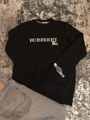Svart tröja från Burberry - Säljer en svart tröja från Burberry med stor vit logga och broderad ryttare på bröstet. Tröjan har rund halsringning, långa ärmar och klassisk passform. Perfekt för dig som vill ha en clean och stilren look med lyxig känsla. Ny ej använd.