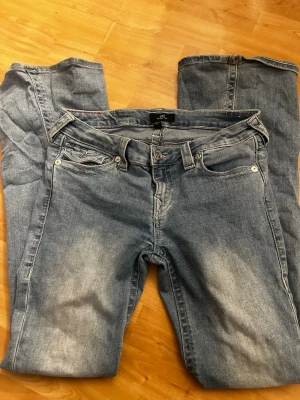 True Religion Joey Low Rise Flare Jeans - Säljer ett par ljusblå True Religion Joey jeans med låg midja och bootcut. Klassiska kontrastsömmar, snygga fickdetaljer med lock och silverknappar bak. Tillverkade i mjukt jeansmaterial, perfekta för dig som gillar en avslappnad men ändå trendig look. Obs Vänstra knappen bak är lite lös.