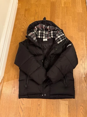 Svart pufferjacka från Burberry - Säljer en svart pufferjacka från Burberry med stor huva och dragkedja. Insidan har ett rutigt foder i svart, vitt och rött. Jackan har två fickor med dragkedja och en tydlig Burberry London England-patch på ärmen. Perfekt för kalla dagar.