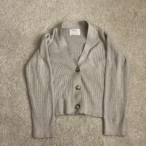 Beige ribbad kofta från Zara - Säljer en beige ribbad kofta från Zara i storlek 152. Koftan har v-ringning, tre stora knappar framtill och långa ärmar. Perfekt att slänga över en topp för en chill look. Materialet är mjukt och stretchigt, vilket gör den superbekväm.