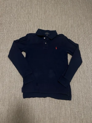 Mörkblå långärmad Polo Ralph Lauren - Klassisk mörkblå långärmad skjorta från Polo Ralph Lauren med krage, två knappar och den ikoniska röda broderade loggan på bröstet. Skjortan är i bomull och har en clean, tidlös look som funkar till det mesta.