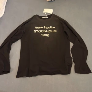 Svart långärmad tröja Acne Studios - Svart långärmad tröja från Acne Studios med vit text 'STOCKHOLM 1996' på bröstet. Klassisk rund hals och mjukt bomullsmaterial. Perfekt för dig som gillar stilrena och trendiga plagg med tydlig logga framtill.