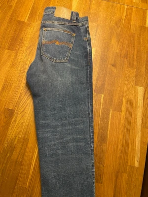 Blå jeans från Nudie Jeans - Säljer ett par klassiska blå jeans från Nudie Jeans storlek W31 L30 med snygga slitningar och kontrastsömmar. Modellen har fem fickor, orange broderi på bakfickorna och normal passform. Tillverkade i slitstarkt bomullstyg med en cool tvättad look. Aldrig använda 