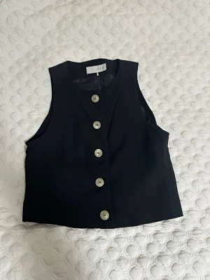 Svart väst med knappar från Zara - Säljer en stilren svart väst från Zara i storlek S. Västen har en rak passform och är försedd med fem silvriga knappar framtill. Perfekt att styla över en skjorta eller t-shirt för en trendig look.