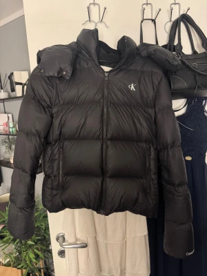 Svart pufferjacka Calvin Klein Jeans XS - Svart pufferjacka från Calvin Klein Jeans i storlek XS. Jackan har en hög krage, avtagbar huva och två fickor med dragkedja. CK-logga på bröstet och Calvin Klein-brodyr på ärmen. Perfekt för kalla dagar med sin fluffiga och stilrena look.