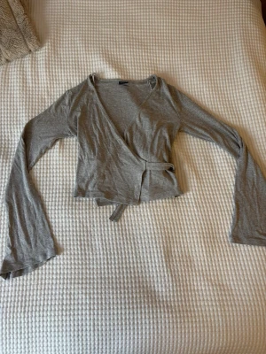 Grå omlottopp med knytband - Säljer en grå omlottopp från Gina Tricot med långa ärmar och knytband i midjan. Toppen har en v-ringad skärning och är tillverkad i mjuk bomullsmix. 