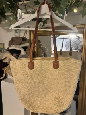Beige shoppingväska med bruna handtag - Snygg beige shoppingväska i flätad stil med långa bruna handtag i skinnimitation. Väskan är rymlig och har dragkedja upptill samt innerfack med dragkedja. Perfekt för strandhäng eller cityrundor.
