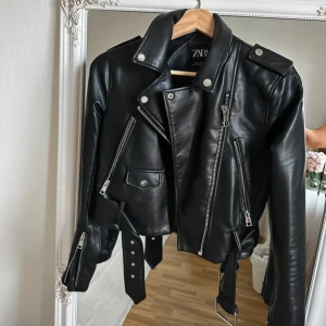 Svart skinnjacka från Zara - Säljer en svart skinnjacka från Zara. Jackan har silvriga dragkedjor, axelklaffar, bälte i midjan och flera fickor med detaljer. 