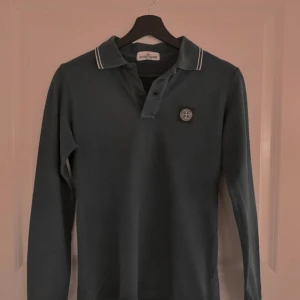 Mörkblå långärmad piké Stone Island - Säljer en mörkblå långärmad pikétröja från Stone Island med klassisk krage och vita ränder på kragen. Tröjan har knappar framtill och den ikoniska Stone Island-loggan på bröstet. Perfekt för dig som gillar stilrena och sportiga plagg.