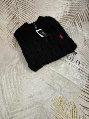 Svart kabelstickad tröja Polo Ralph Lauren - Kabelstickad svart tröja från Polo Ralph Lauren med halv dragkedja och röd broderad logga på bröstet. Tröjan har ribbade muddar och hög krage, perfekt för kyliga dagar. Tillverkad i mjuk merinoull för extra komfort.