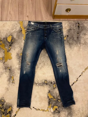 Replay blå jeans med slitningar - Säljer ett par snygga Replay jeans i mörkblå tvätt med slitna detaljer och knappar i gylfen. Modellen har klassiska fem fickor och coola slitningar på låren och knäna. Jeansen har en smal passform och är tillverkade i jeansmaterial.