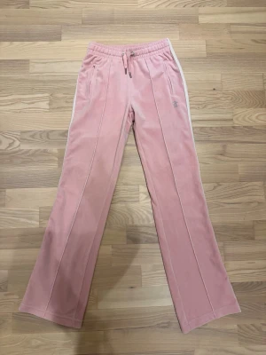 Rosa Juicy Couture Set - Säljer ett par klassiska rosa mjukisbyxor och zip tröja från Juicy Couture i mjuk velour. Byxorna har normal passform, raka ben och resårmidja med snörning. Sidorna har vita detaljer och på baksidan står det 'Juicy' i glittriga stenar. De är äkta och har bara används 2 gånger. Pris går att diskutera 