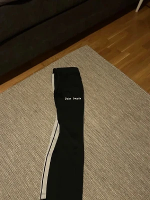 Palm Angels svarta träningsbyxor - Svarta träningsbyxor från Palm Angels med vita sidoränder och logga på låret. Byxorna har en rak passform och elastisk midja för skön komfort. Perfekta för en sportig och trendig look. Aldrig använda bara 2,3 gånger, storlek L men känns mer som M