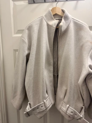 Ljusgrå ulljacka från Zara - Säljer en stilren ljusgrå jacka från Zara i ullmix. Jackan har hög krage, raka axlar och bälte med spänne i nederkant för extra detalj. Perfekt oversized passform och enkel design utan synliga knappar eller dragkedja framtill.