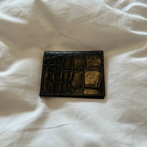 Svart Mulberry plånbok i skinn med krokoprägling - Snygg svart plånbok från Mulberry i äkta skinn med krokodilpräglad yta. Insidan har flera kortfack och är fodrad med brunt tyg med Mulberrys klassiska trädmönster. Perfekt för dig som vill ha en stilren och exklusiv accessoar.