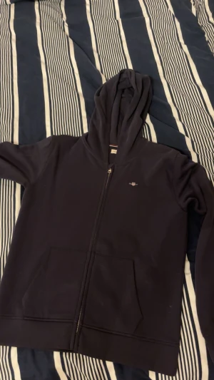 Navy blue hoodie från GANT med zip - Navy blue hoodie från GANT med broderad logga på bröstet och praktisk huva. Tröjan har hel dragkedja framtill, två stora fickor och ribbade muddar. Insidan har en snygg randig detalj vid halsen. Perfekt för chill eller häng med kompisar. Detta är något för dig som vill ha en stilren look men samtidigt inte skrika märkeskläder. Tröjan är använd endast 1 gång. Pris går att diskuteras vid snabb affär