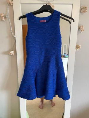  kortklänning från Aqua - Säljer en snygg blå kortklänning från Aqua. Klänningen är ärmlös med rund halsringning och har ett vågigt ribbat mönster i tyget. Kjolen är utställd och materialet känns som polyester med ett mjukt foder. Perfekt för dig som vill sticka ut!