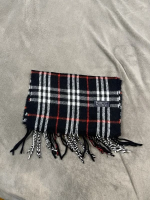 Rutig halsduk från Burberrys i ull - Säljer en klassisk rutig halsduk från Burberrys i 100% lammull. Halsduken har svart bas med vita och röda rutor samt tvinnade fransar i ändarna. Märkt med Burberrys of London, Made in England. Perfekt accessoar för kyliga dagar. P8