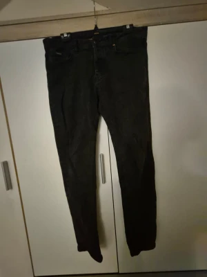 Svarta jeans från BOSS - Säljer ett par svarta jeans från BOSS med klassisk femficksdesign och snygga orangea detaljer på insidan och bakfickan. Jeansen har rak passform och är tillverkade i ett slitstarkt bomullsmaterial. Perfekta för dig som gillar stilrena och tidlösa plagg.