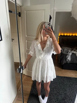 Vit kort klänning med spets och volang - Supersöt vit kort klänning med broderade spetsdetaljer, omlott-v-ringning och volangkant. Armarna har knappar så man kan stänga, se bild 3. Och en dragkedja på vänster sidan för att enklare få på sig den. Står strl 38 och den passar mig som brukar ha S/M i kläder. Lagom lång på mig som är 168cm. Undra du nått är de bara att skriva 