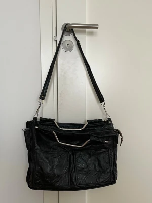 Vintage axelväska i skinn - Säljer en svart axelväska i skinn med två stora ytterfickor framtill och avtagbar axelrem. Väskan har metallhandtag och dragkedja upptill. Perfekt för dig som vill ha plats för allt och gillar en stilren look. VINTAGE!!❤️