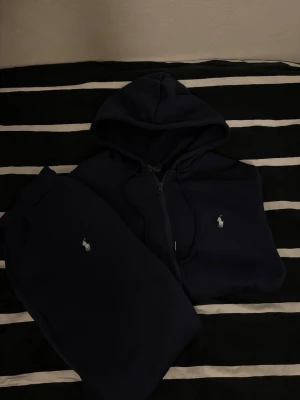 Mörkblå mjukisbyxor och hoodie från Polo Ralph Lauren - Säljer ett par mörkblå mjukisbyxor och hoodie från Polo Ralph Lauren med klassisk vit logobrodyr på benet. Byxorna har ribbade muddar och är gjorda i mjukt bomullsmaterial, perfekta för chill dagar. Snygg och enkel design med skön passform. I nyskick, hör av dig om du har några frågor.