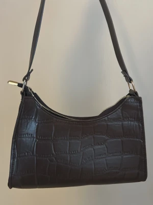 Brun axelväska i krokopräglat skinn - Säljer en mörkbrun axelväska i krokopräglat skinn med stilren form och silverfärgad dragkedja. Väskan har ett justerbart axelband och en elegant look som passar till många outfits.🤎