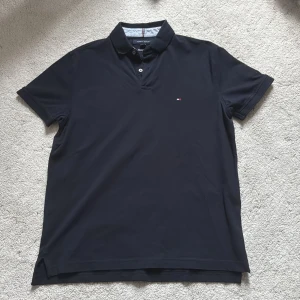 Blå pikétröja från Tommy Hilfiger - Klassisk blå pikétröja från Tommy Hilfiger med liten broderad logga på bröstet. Tröjan har krage, två knappar vid halsen och korta ärmar. Tillverkad i mjuk bomull som känns skön mot huden. Perfekt till jeans eller shorts.