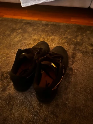 Nike Air Force 1 svart/gul sneakers - Säljer ett par svarta Nike Air Force 1 sneakers med gula detaljer och logga. Skorna har klassisk snörning, perforerad tå och en låg siluett. Materialet är en mix av skinn och textil, med svart sula och gul Nike Swoosh-kant. Perfekta för dig som gillar streetwear och stilrena färger.