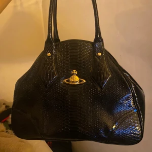 Äkta Vivienne Westwood - Säljer en svart handväska från Vivienne Westwood i veganskt läder med ormmönstrad yta och guldfärgade detaljer. Väskan har dubbla handtag, ikonisk logga framtill och dragkedja med präglad logotyp. Väskan är köpt från Vestiaire Collective med authentication and quality control. Väskan är stor och sparsamt använd från förra ägaren, använd 1 gång av mig. FINNS I GÖTEBORG. Säljer billigt pga att jag inte har kvitto eller liknande från själva märket, men väskan är 100% äkta.