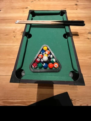 Mini biljardbord  - perfint och smidigt litet biljardbord  perfekt för roliga matcher hemma. Bordet är i bra skick och kommer med allt du behöver: två köer, triangel och kompletta bollar. Passar både barn och vuxna som vill ha kul utan att behöva ett stort bord!  Mått: ca 50 cm på längden och ca 30 på bredd