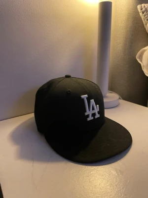 Svart LA-keps från New Era 59FIFTY - Säljer en svart New Era 59FIFTY keps med LA-logga i vitt framtill. Klassisk modell med rak skärm och broderade detaljer. Kepsen är tillverkad i 100% polyester och har MLB-logga bak. Perfekt för dig som gillar streetwear och sportig stil.