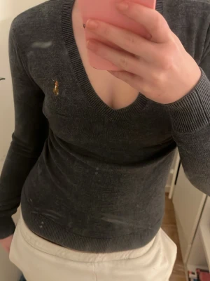 Mörkgrå v-ringad tröja från Ralph Lauren - Säljer en mörkgrå v-ringad tröja från Ralph Lauren med den klassiska broderade polospelaren på bröstet. Tröjan är långärmad och har ribbade muddar vid ärmslut och nederkant. Perfekt för dig som gillar en stilren och tidlös look.