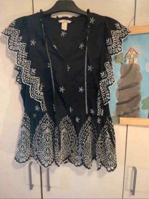 Svart blus med vita broderade detaljer - Supersnygg svart blus med vita broderade mönster och volangkantade ärmar. Blusen har v-ringning och fina spetsdetaljer längs kanterna samt ett unikt broderat mönster längst ner. PM för mer bilder eller vid frågor!💓