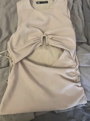 Beige omlottklänning från Zara - Säljer en beige omlottklänning från Zara med snygg snörningsdetalj framtill och öppen midja. Klänningen har en kort längd och är tillverkad i ett mjukt syntetmaterial. Perfekt för dig som gillar trendiga och stilrena plagg med unika detaljer.