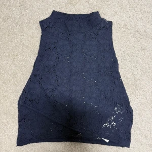 Mörkblå spetstopp från Hollister - Säljer en mörkblå ärmlös croptop från Hollister i storlek S. Toppen har hög hals och är helt täckt av ett vackert spetsmönster som ger en elegant och luftig känsla. Perfekt att styla med jeans eller kjol för en trendig look.