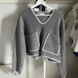 Grå stickad tröja med fickor från Zara - Grå stickad tröja från Zara med två stora framfickor och vit kant runt hals, ärmslut och nederkant. Tröjan har v-ringning 