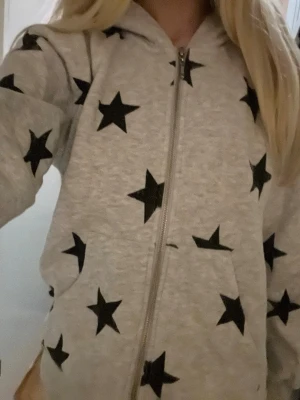 Zip up hoddie - Stilren och bekväm zip-up hoodie i grå färg med coola svarta stjärndetaljer. Tillverkad i mjukt och skönt material som passar perfekt för både vardag och fritid. Dragkedja framtill gör den enkel att ta av och på, medan huvan ger extra komfort och en avslappnad look. En modern hoodie som sticker ut och passar lika bra till jeans som till mjukisbyxor.
