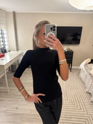 Svart ribbad topp med halvärm - Säljer en svart ribbad topp med halvärm och högre hals. Toppen är figurnära och har en stilren look som passar till mycket. Materialet är stretchigt och bekvämt, perfekt för dig som gillar en enkel men snygg stil.