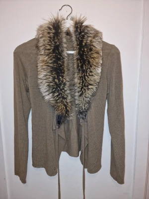 Beige kofta med fuskpäls och knytband - Beige/ljusbrun kofta med stor, fluffig fuskpäls runt kragen och långa ärmar. Koftan har ett knytband i midjan för snygg passform och är tillverkad i ett mjukt stickat material. Perfekt för dig som vill ha en mysig och trendig look i höst och vinter.